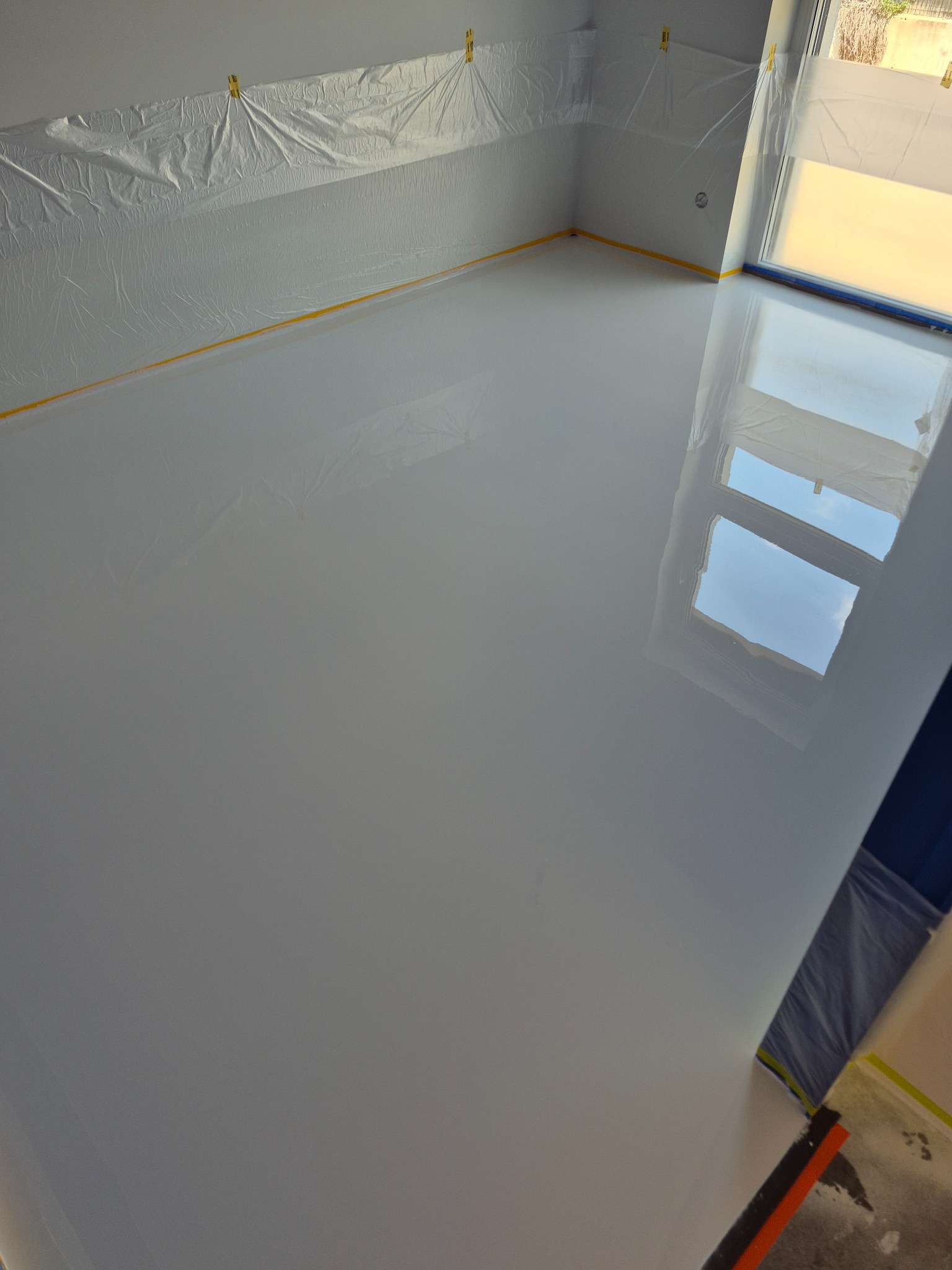 Biała, lśniąca posadzka w pokoju, zabezpieczone ściany folią malarską. Widoczne odbicie okna w gładkiej powierzchni podłogi.