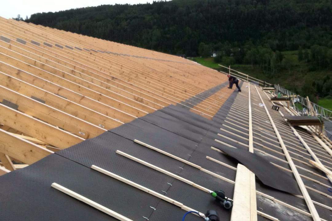 Konstrukcja dachu z drewnianymi belkami, pokryta czarną membraną hydroizolacyjną, częściowo jeszcze niezakryta, widoczny pracownik i narzędzia pneumatyczne.