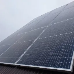 Panel fotowoltaiczny na brązowym, falistym dachu, zamontowany przy pomocy srebrnych uchwytów, widok pod kątem w pochmurny dzień.