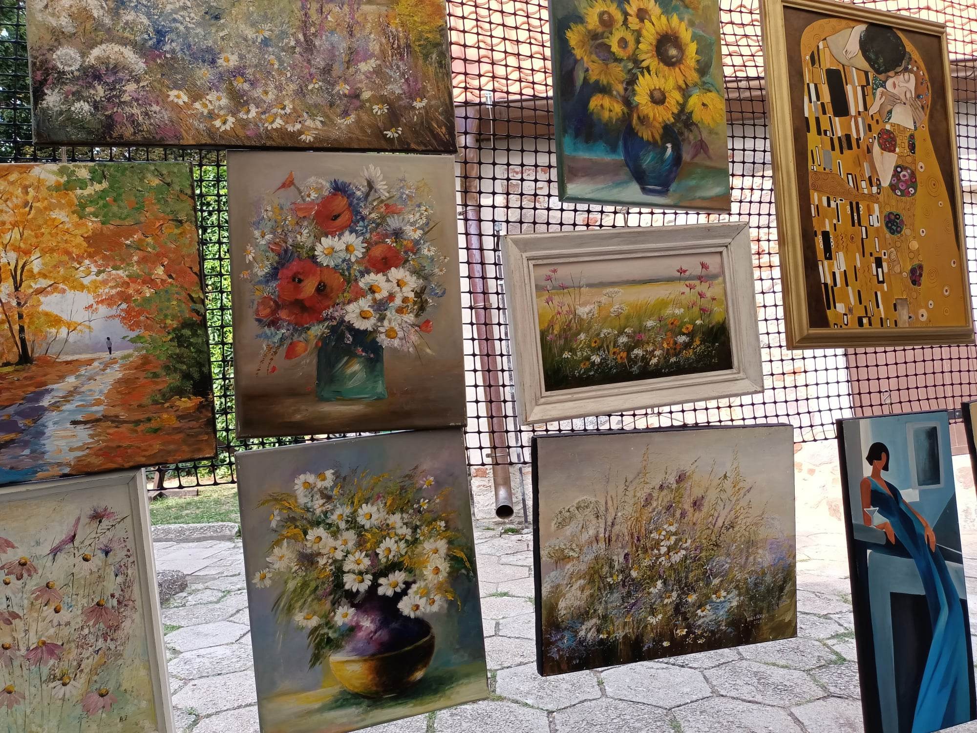 Różnorodne obrazy olejne, w tym kwiaty, pejzaże i reprodukcja 'Pocałunku' Gustava Klimta, prezentowane na zewnątrz, na tle brukowanej nawierzchni.