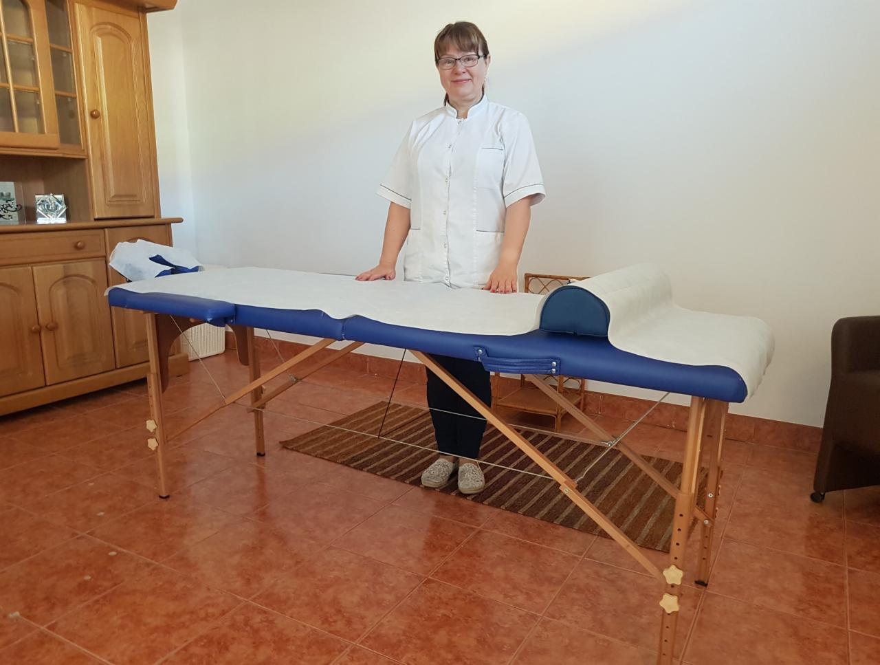 Kobieta w białym uniformie opiera ręce na niebieskim stole do masażu z drewnianą ramą, przygotowanym do zabiegu, na brązowej podłodze z terakoty.