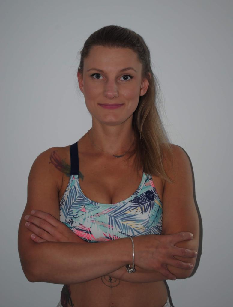 Kobieta w sportowym topie z motywem tropikalnych liści, skrzyżowanymi rękami, tatuażem na ramieniu i srebrną bransoletką, na tle jasnej ściany.