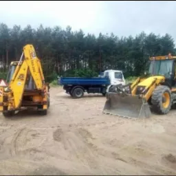 Dwie ż&oacute;łte koparko-ładowarki JCB i ciężar&oacute;wka z niebieską skrzynią ładunkową na piaszczystym terenie z lasem w tle.