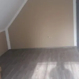 Pokój na poddaszu po remoncie: beżowe ściany z białymi listwami, szare panele podłogowe z widocznym wzorem drewna, skos dachu.