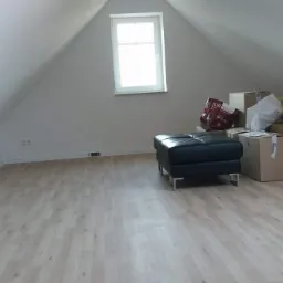 Poddasze z nową, jasną podłogą panelową, widoczne kartony i pufa, białe ściany i okno w szczycie dachu.