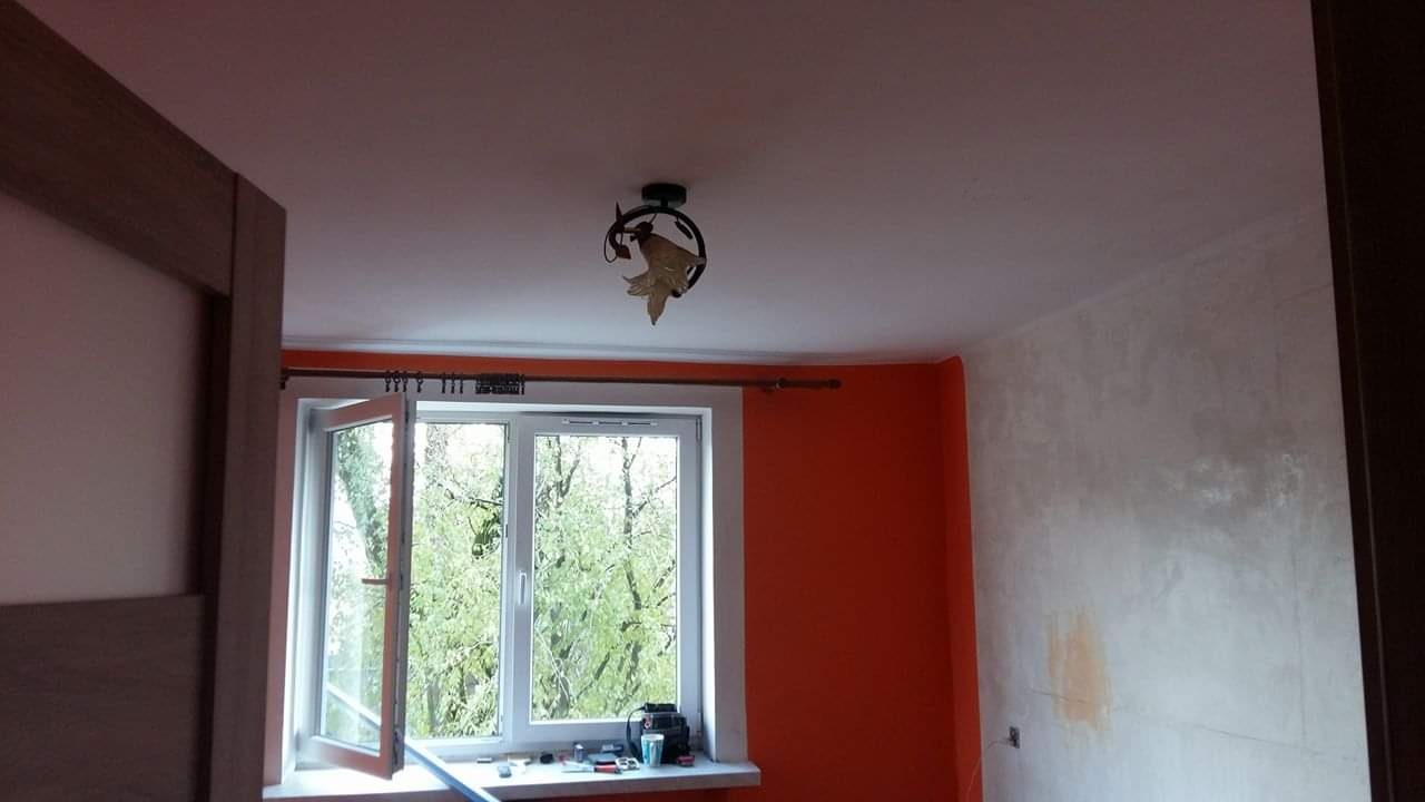 Pokój w trakcie remontu: pomarańczowa ściana kontrastuje z surową, szpachlowaną powierzchnią, okno otwarte na zieleń, lampa z motywem liści na suficie.
