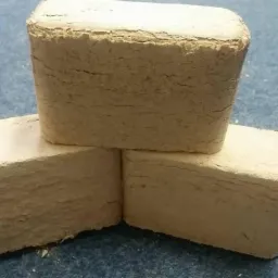 Trzy kostki sprasowanego materiału opałowego ułożone jedna na drugiej na szaro-niebieskim tle, widoczne warstwy materiału i drobne pęknięcia na powierzchni.