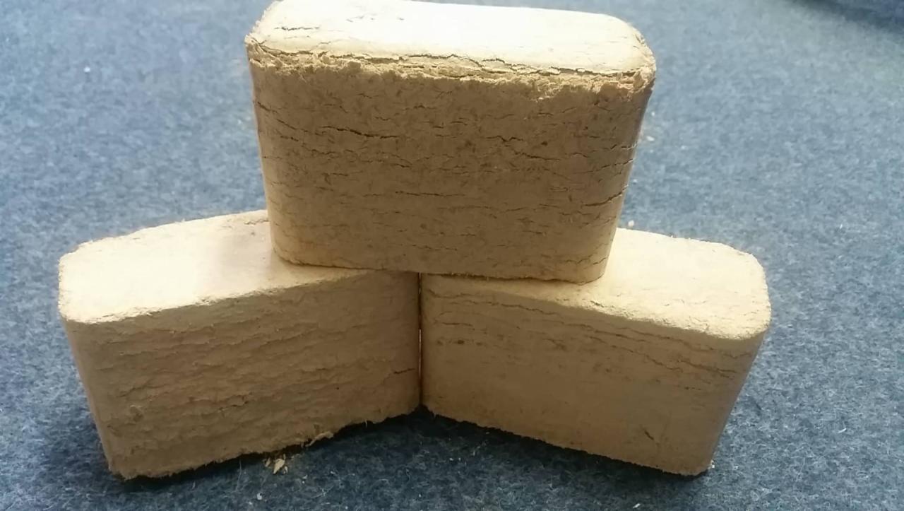 Trzy kostki sprasowanego materiału opałowego ułożone jedna na drugiej na szaro-niebieskim tle, widoczne warstwy materiału i drobne pęknięcia na powierzchni.