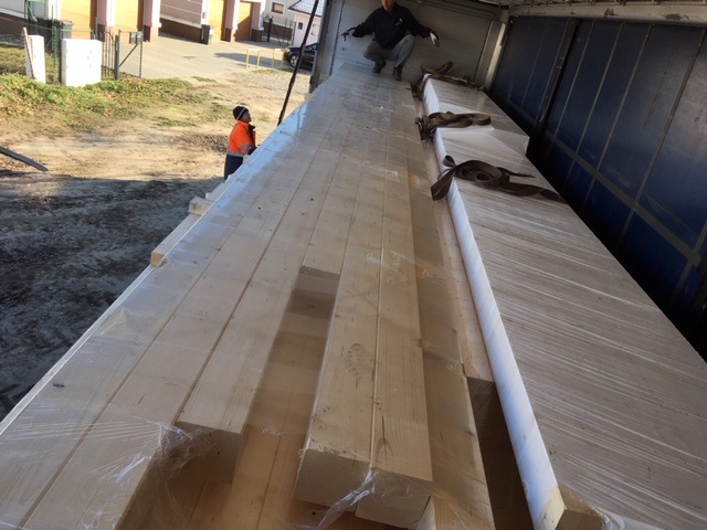 Ładunek tarcicy budowlanej w folii ochronnej, zabezpieczony pasami transportowymi wewnątrz naczepy, widoczny pracownik w odblaskowej kamizelce.