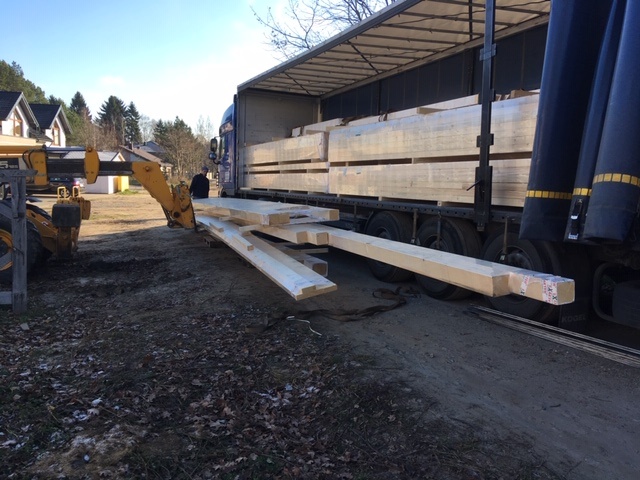 Żółty wózek widłowy rozładowuje pakiet jasnej tarcicy z ciężarówki z naczepą na placu budowy.