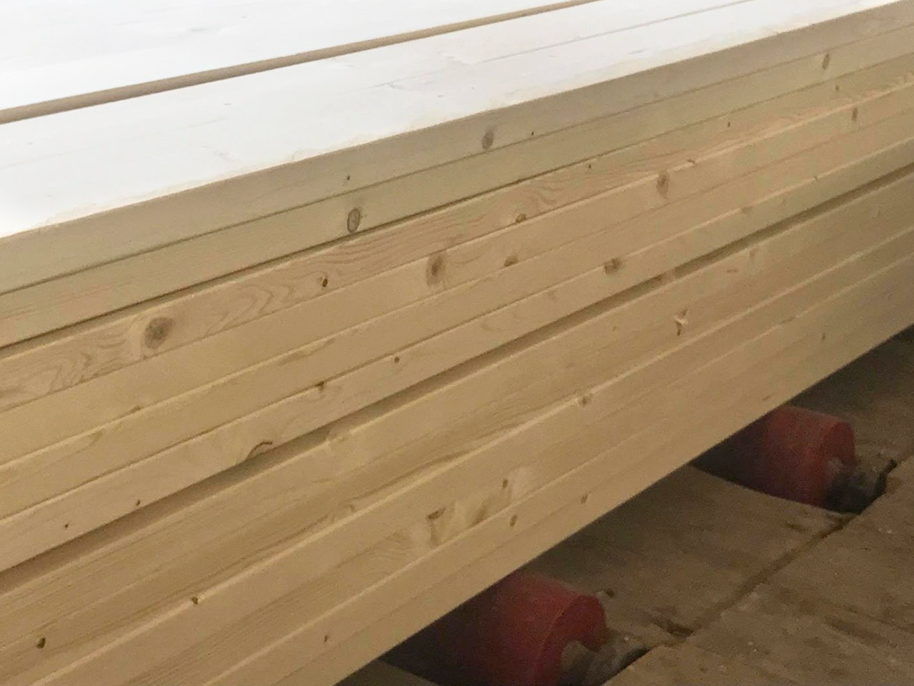 Stos jasnej, świeżo obrobionej tarcicy ułożonej na czerwonych rolkach transportowych w magazynie.