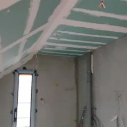 Skośny sufit na poddaszu z zamontowaną płytą gipsowo-kartonową, widoczne szpachlowane łączenia, okno w metalowej ramie, przewody elektryczne wystające ze ściany, szara rura kanalizacyjna przy ścianie.