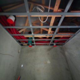 Edil Constration Bud - Instalacja sufitowa z czerwonymi rurami w surowym wnętrzu, widoczne drewniane elementy konstrukcyjne i metalowa rama.
