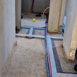 Edil Constration Bud - Instalacja wodna w trakcie montażu: czerwone i niebieskie rury prowadzone wzdłuż metalowej konstrukcji w surowym wnętrzu z oknem w tle.