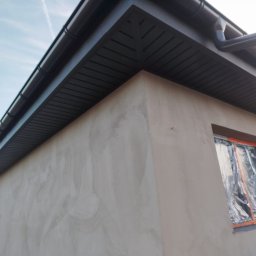 Edil Construction BUD Andrzej Godlewski - Fragment budynku po nałożeniu szarego tynku, z widoczną czarną podbitką dachową i oknem zabezpieczonym folią. Ujęcie z dołu podkreśla geometrię budynku.