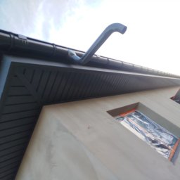 Edil Construction BUD Andrzej Godlewski - Szara elewacja domu z zamontowanym systemem rynnowym, okno zabezpieczone folią. Widok z dołu, podkreślający detale wykończenia i przygotowanie do dalszych prac.