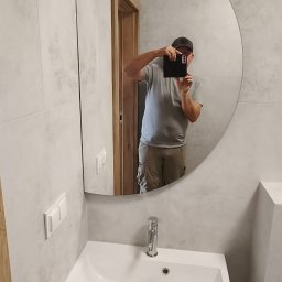 Edil Construction BUD Andrzej Godlewski - Nowoczesna łazienka z umywalką w szafce i okrągłym lustrem, w którym odbija się osoba robiąca zdjęcie. Ściany wykończone płytkami imitującymi beton.
