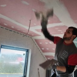 Edil Construction BUD Andrzej Godlewski - Gipsowanie sufitu z płyt karton-gips przez pracownika w trakcie remontu poddasza. Widoczne łączenia płyt i szpachlowane fragmenty. Okno w tle.