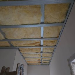 Edil Construction BUD Andrzej Godlewski - Sufit podwieszany z metalową konstrukcją i izolacją z wełny mineralnej, widok z dołu, drabina oparta o ścianę, surowy stan wykończenia wnętrza.
