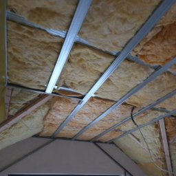 Edil Construction BUD Andrzej Godlewski - Ocieplenie poddasza wełną mineralną, widoczne metalowe profile konstrukcyjne i drewniane elementy więźby dachowej. Prace wykończeniowe w trakcie realizacji.