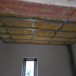 Edil Construction BUD Andrzej Godlewski - Sufit podwieszany z izolacją z wełny mineralnej, widok z dołu na metalową konstrukcję i ocieplenie, w tle okno i surowa ściana. Etap prac wykończeniowych.