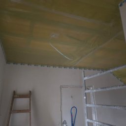 Edil Construction BUD Andrzej Godlewski - Montaż izolacji na suficie podwieszanym z widocznymi przewodami elektrycznymi, drabina i rusztowanie w surowym wnętrzu. Etap prac wykończeniowych.