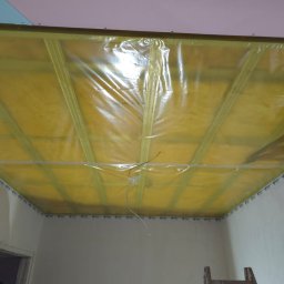 Edil Construction BUD Andrzej Godlewski - Ocieplenie poddasza z widoczną folią paroizolacyjną i stelażem pod płyty gipsowo-kartonowe. Widoczna drabina i fragment osoby w dolnym rogu.
