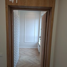 Edil Constration Bud - Otwarty widok na wnętrze mieszkania z parkietem w jodełkę, białą ścianą z dekoracyjną ramą i ażurową ścianką działową. Numer '41' nad wejściem.
