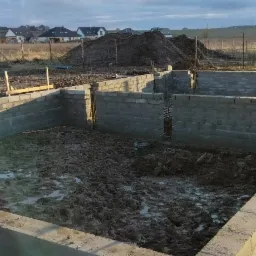 Wznoszenie fundamentów z bloczków betonowych; widoczne zbrojenia, niebieski wąż i butelka napoju na murze; w tle domy jednorodzinne i hałda ziemi.