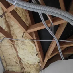 Izolacja poddasza wełną mineralną, widoczne drewniane krokwie, rury instalacyjne i fragment okna dachowego.