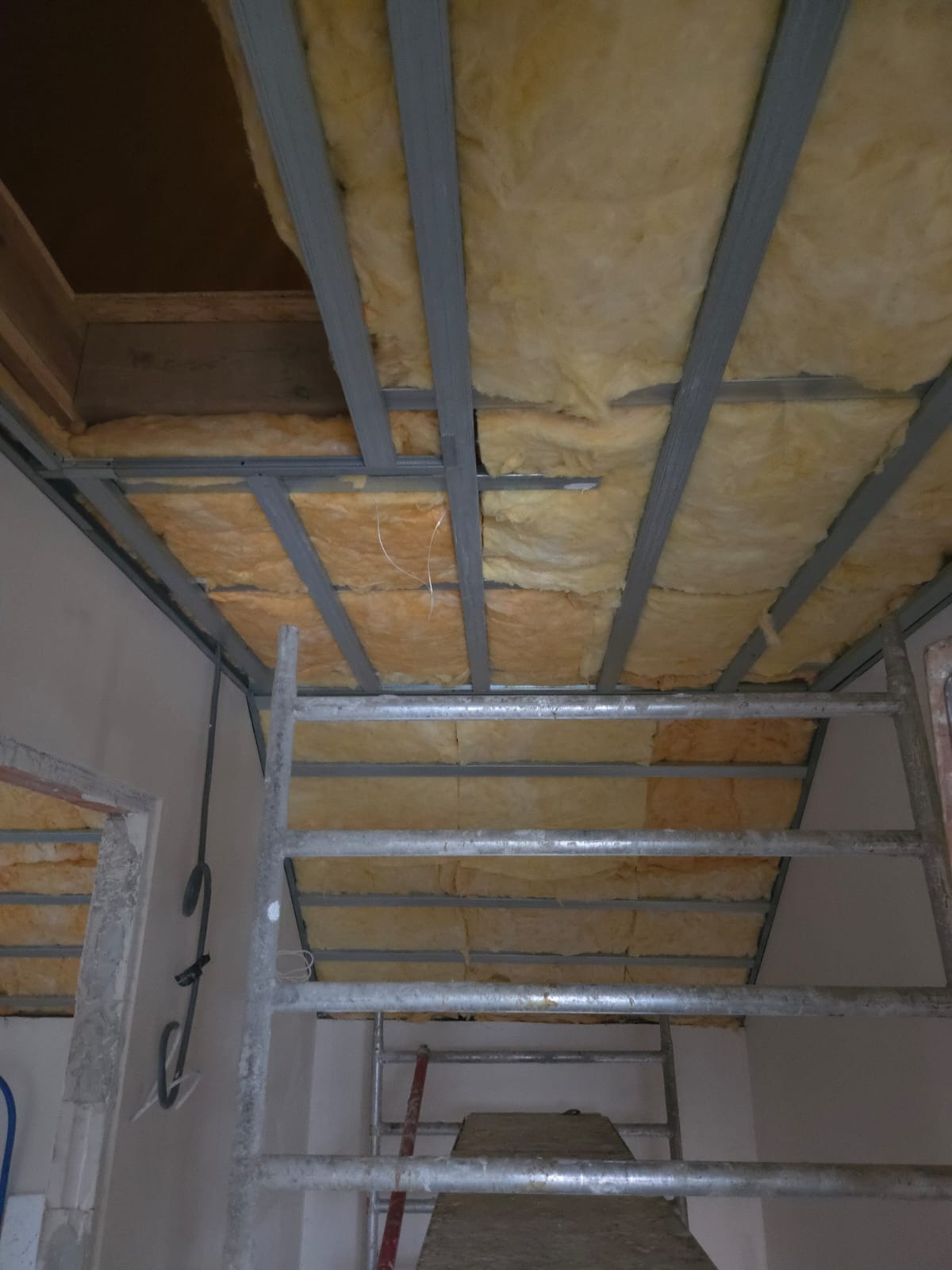 Montaż izolacji z wełny mineralnej w metalowym stelażu sufitu podwieszanego, widok z dołu, drabina w kadrze. Prace wykończeniowe w trakcie realizacji.