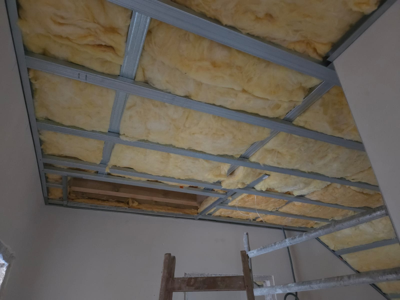 Montaż izolacji z wełny mineralnej w metalowym stelażu sufitu podwieszanego. Widoczne fragmenty drabiny malarskiej.