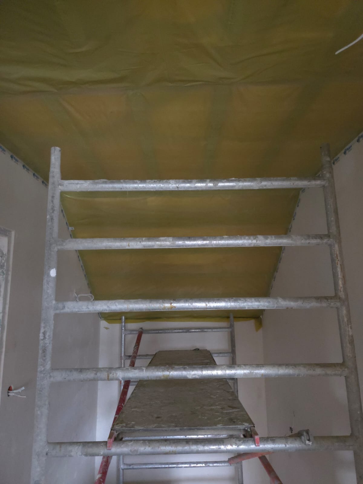 Sufit zabezpieczony folią budowlaną, widok ze rusztowania. Przygotowanie do prac wykończeniowych, widoczne ślady po taśmie malarskiej na ścianach.