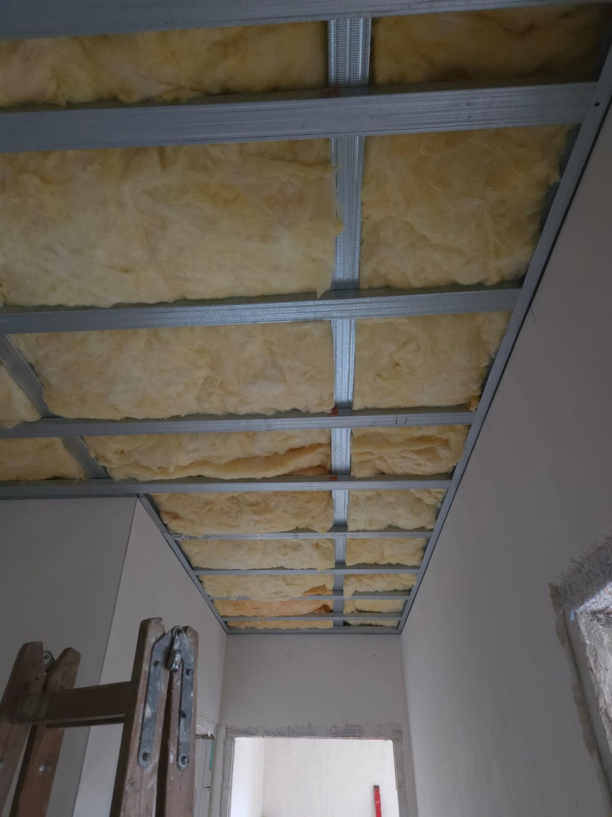 Sufit podwieszany z metalową konstrukcją i izolacją z wełny mineralnej, widok z dołu, drabina oparta o ścianę, surowy stan wykończenia wnętrza.