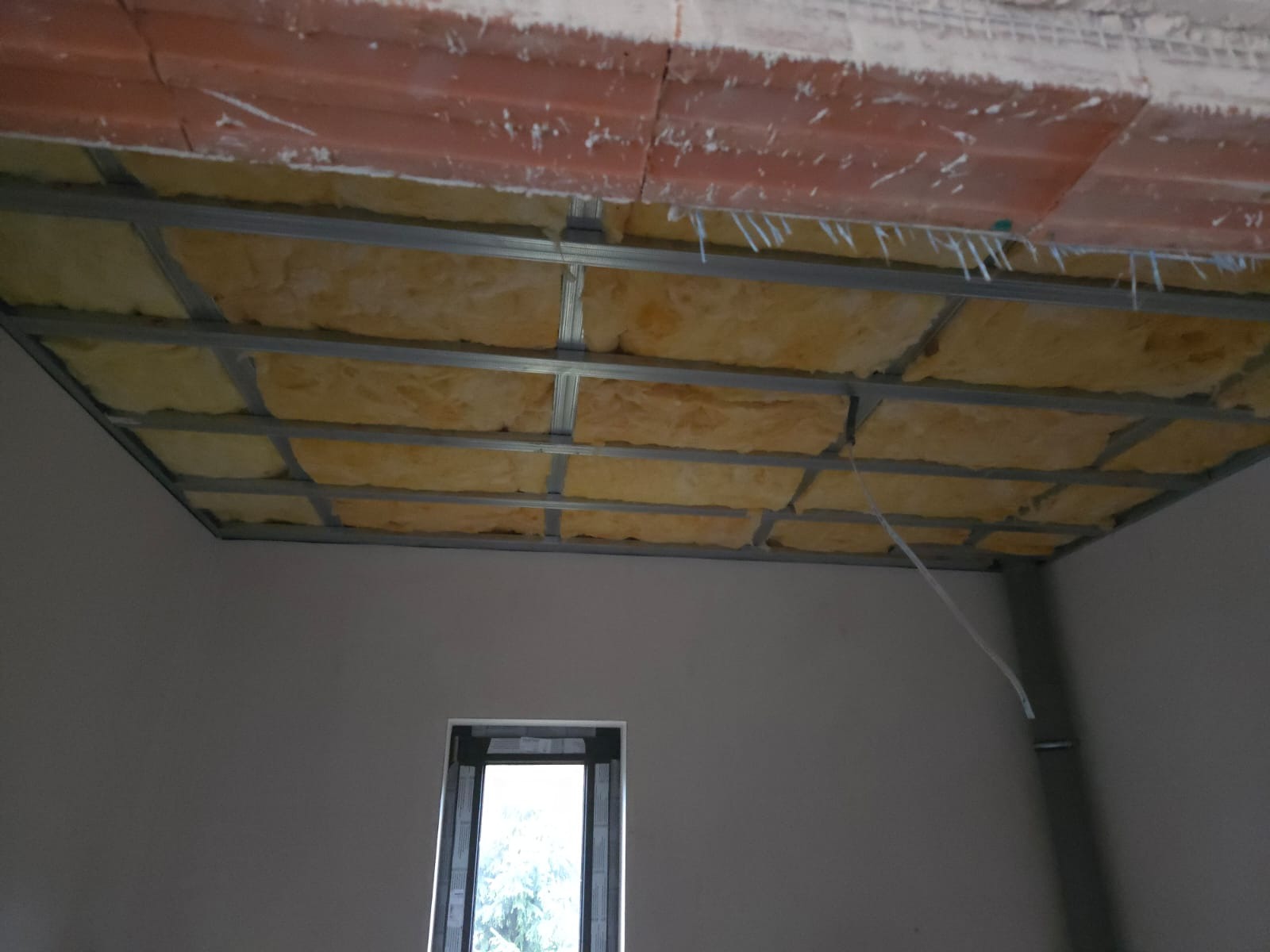 Sufit podwieszany z izolacją z wełny mineralnej, widok z dołu na metalową konstrukcję i ocieplenie, w tle okno i surowa ściana. Etap prac wykończeniowych.