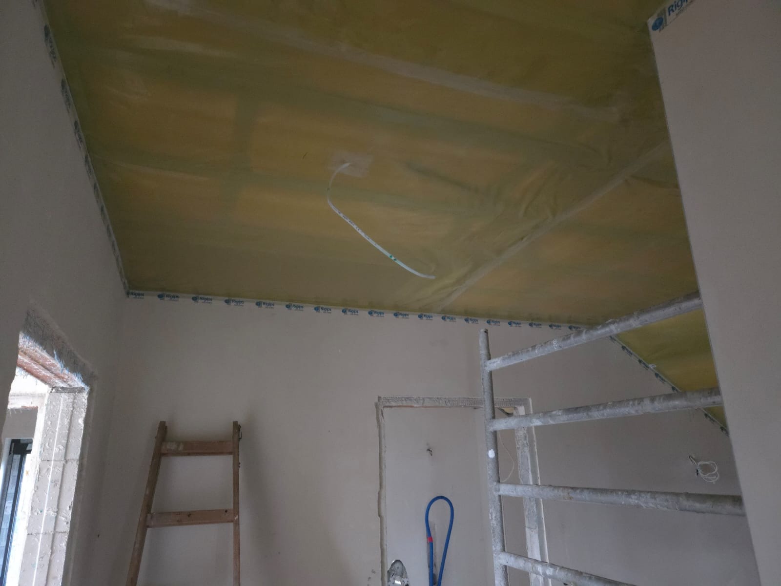 Montaż izolacji na suficie podwieszanym z widocznymi przewodami elektrycznymi, drabina i rusztowanie w surowym wnętrzu. Etap prac wykończeniowych.