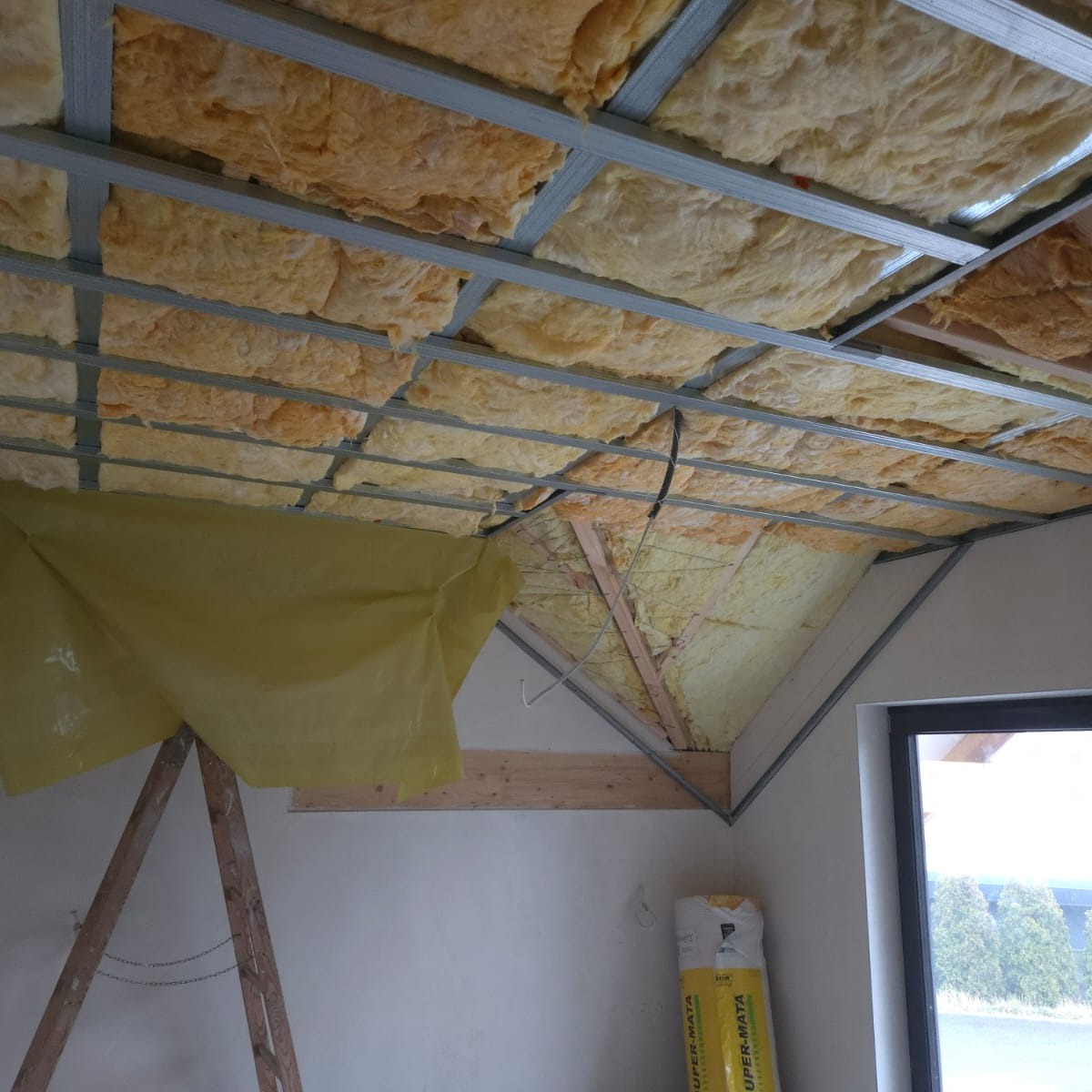 Ocieplenie poddasza wełną mineralną, widoczne metalowe profile, folia ochronna i fragment okna. Prace wykończeniowe w trakcie realizacji.