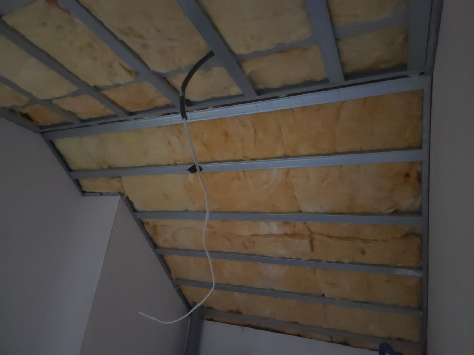 Sufit podwieszany z izolacją z wełny mineralnej, widoczne profile metalowe i przewody elektryczne. Kąt ujęcia od dołu, oświetlenie naturalne.