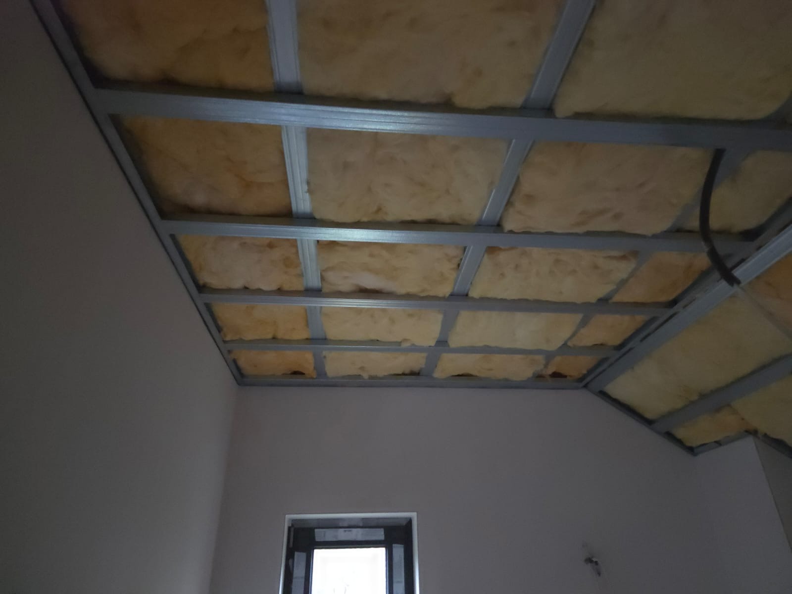 Sufit podwieszany z izolacją z wełny mineralnej, widoczne metalowe profile. Prace wykończeniowe w pomieszczeniu z oknem. Ujęcie z dołu, perspektywa wnętrza.