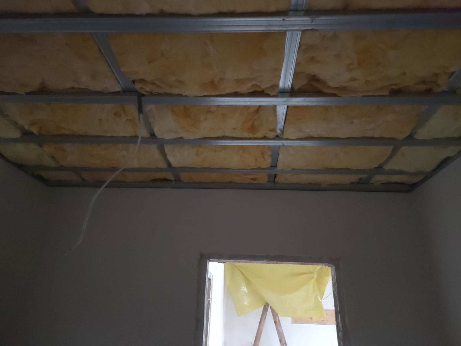 Sufit podwieszany z izolacją z wełny mineralnej, widoczne profile metalowe i fragment okna zabezpieczonego folią malarską. Przewód elektryczny zwisa z sufitu.