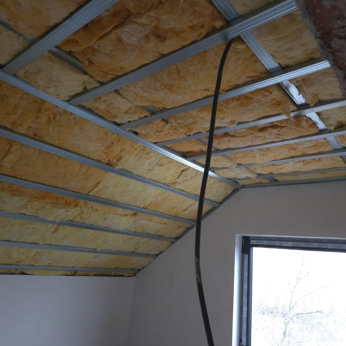 Ocieplenie poddasza wełną mineralną, widoczne metalowe profile konstrukcyjne sufitu podwieszanego i fragment okna. Przewód wentylacyjny zwisa z sufitu.