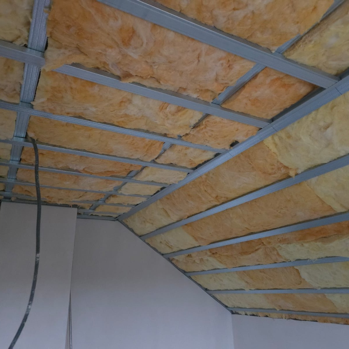 Sufit podwieszany z izolacją z wełny mineralnej, widoczne profile metalowe i fragment ściany. Prace wykończeniowe na poddaszu.