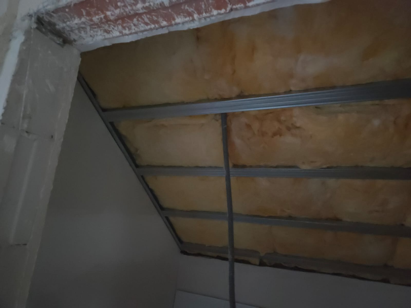 Skośny sufit poddasza z izolacją z wełny mineralnej, widoczne metalowe profile konstrukcyjne. Ujęcie z dołu, fragment ściany w lewym dolnym rogu.