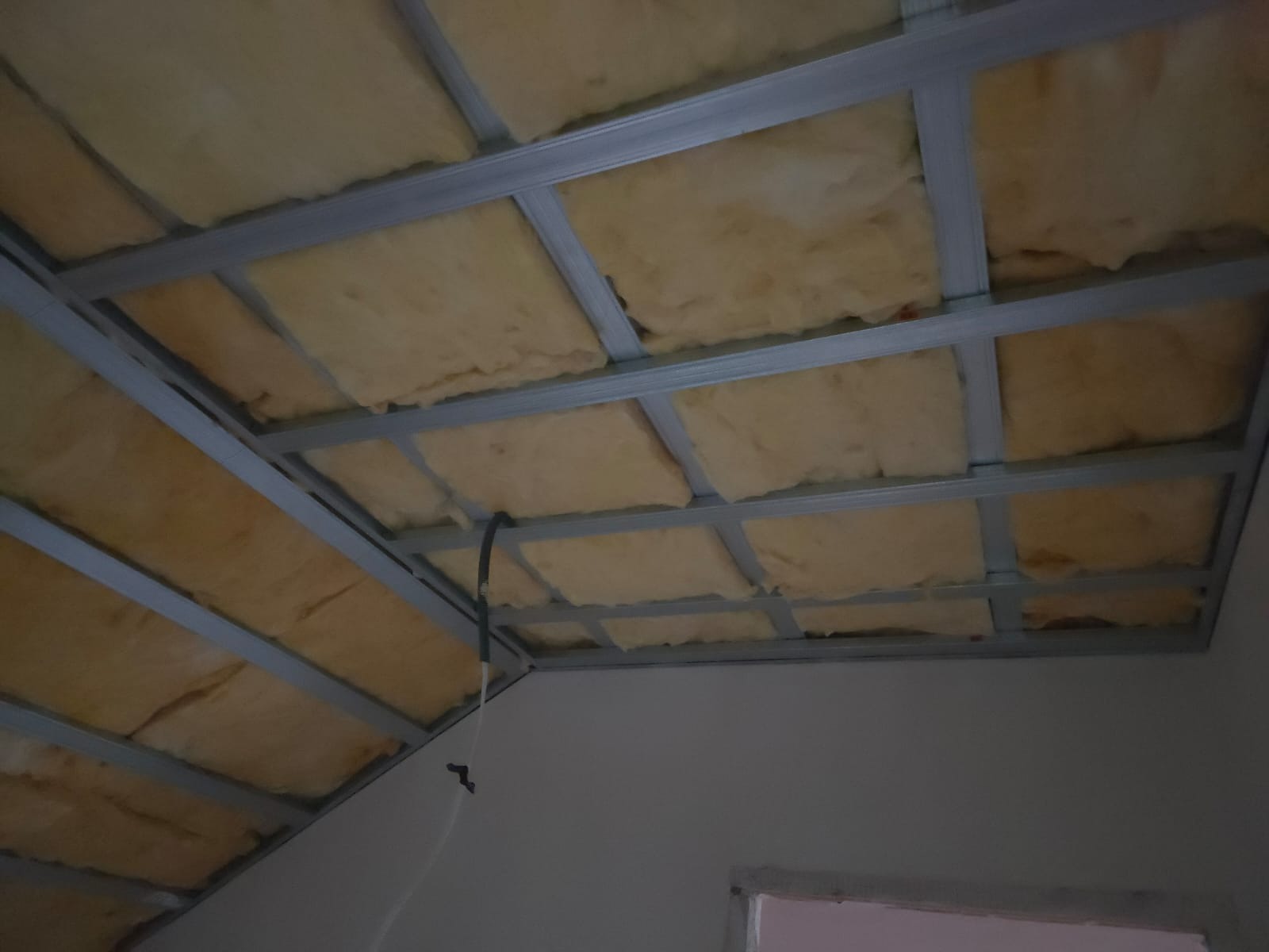 Sufit podwieszany z izolacją z wełny mineralnej, widok z dołu. Metalowa konstrukcja kratowa. Kabel elektryczny zwisający z sufitu. Etap prac wykończeniowych.
