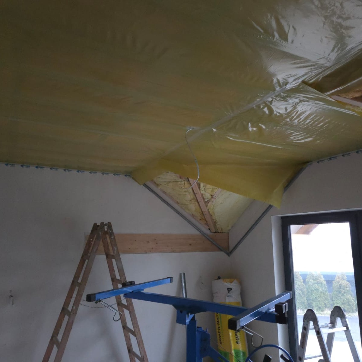 Montaż folii paroizolacyjnej na poddaszu, widoczne profile, izolacja wełną mineralną, podnośnik do płyt i drabina. Prace wykończeniowe na ukończeniu.