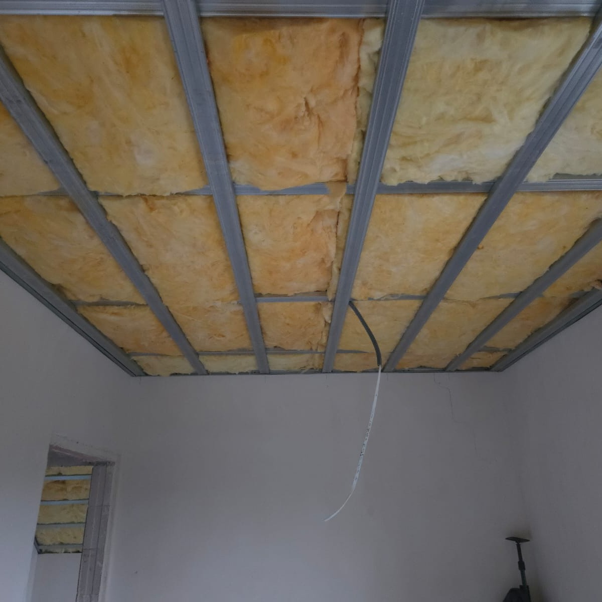 Sufit podwieszany z izolacją z wełny mineralnej, widoczne metalowe profile i fragment ściany. Przewód elektryczny zwisa z sufitu. Izolacja termiczna w trakcie montażu.
