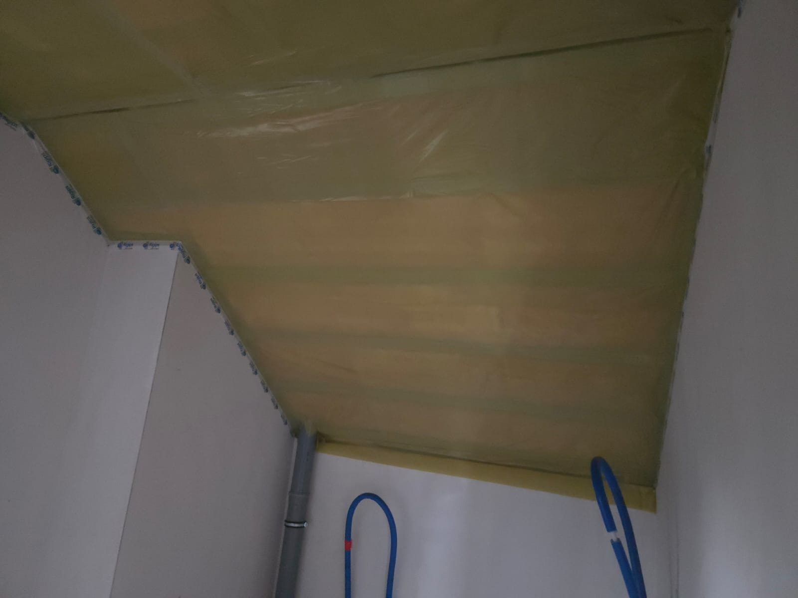 Ocieplenie poddasza wełną mineralną, zabezpieczone folią. Widoczne rury instalacyjne przy ścianie. Ukośny sufit z izolacją termiczną w trakcie prac wykończeniowych.