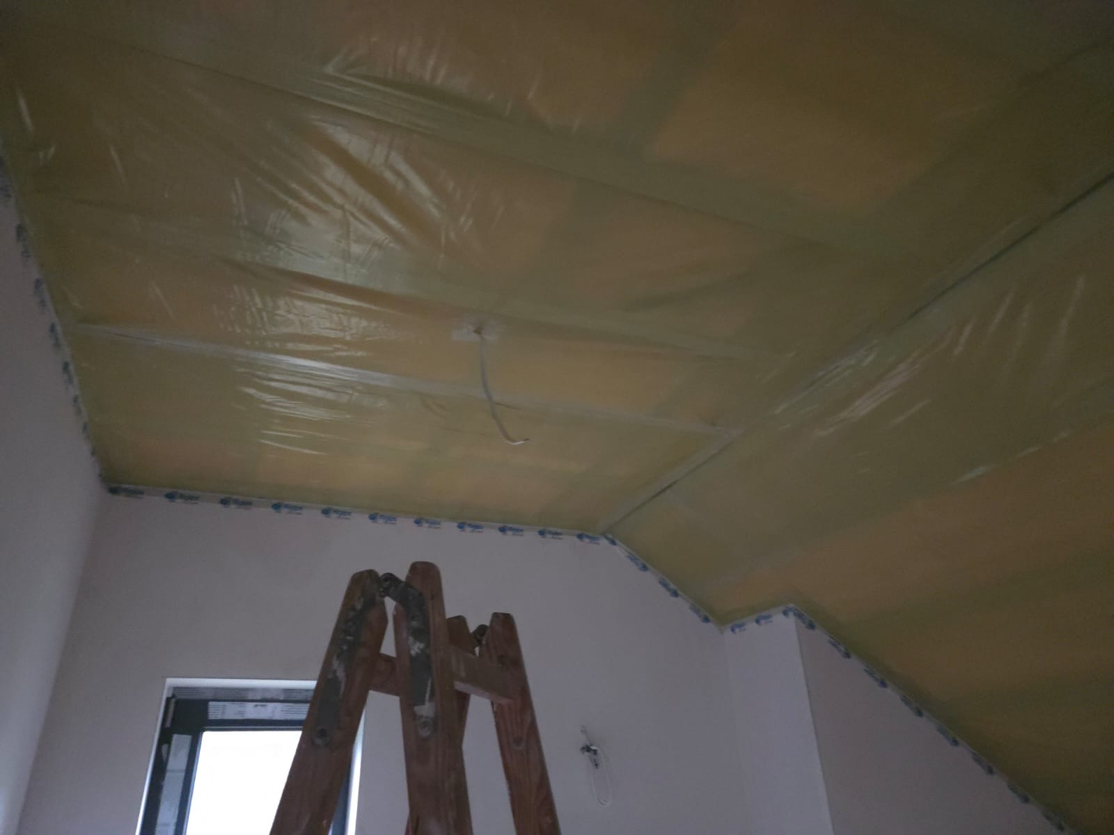 Skośny sufit zabezpieczony folią malarską, widoczna drabina malarska i fragment okna. Przygotowanie do malowania lub montażu płyt karton-gips.