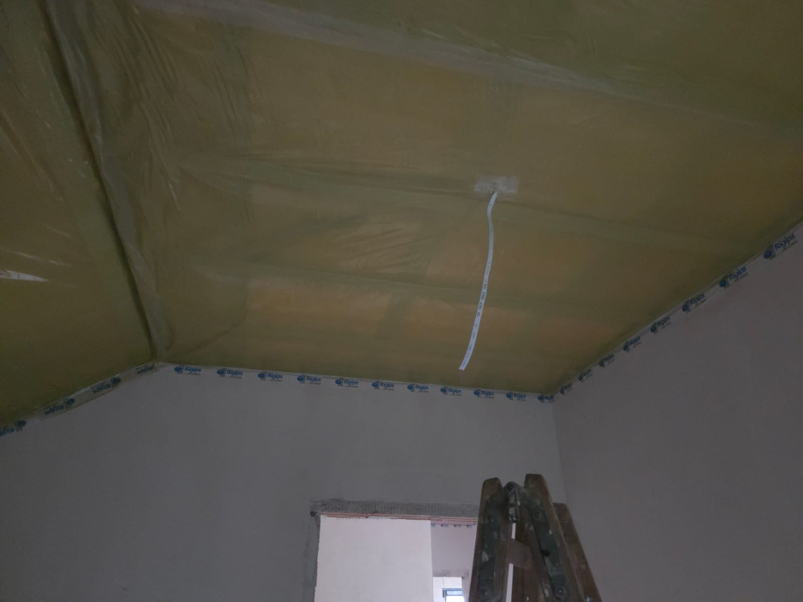 Skośny sufit zabezpieczony folią i taśmą Rigips, z wystającym przewodem elektrycznym. W tle fragment drabiny i otwór drzwiowy. Przygotowanie do montażu płyt karton-gips.