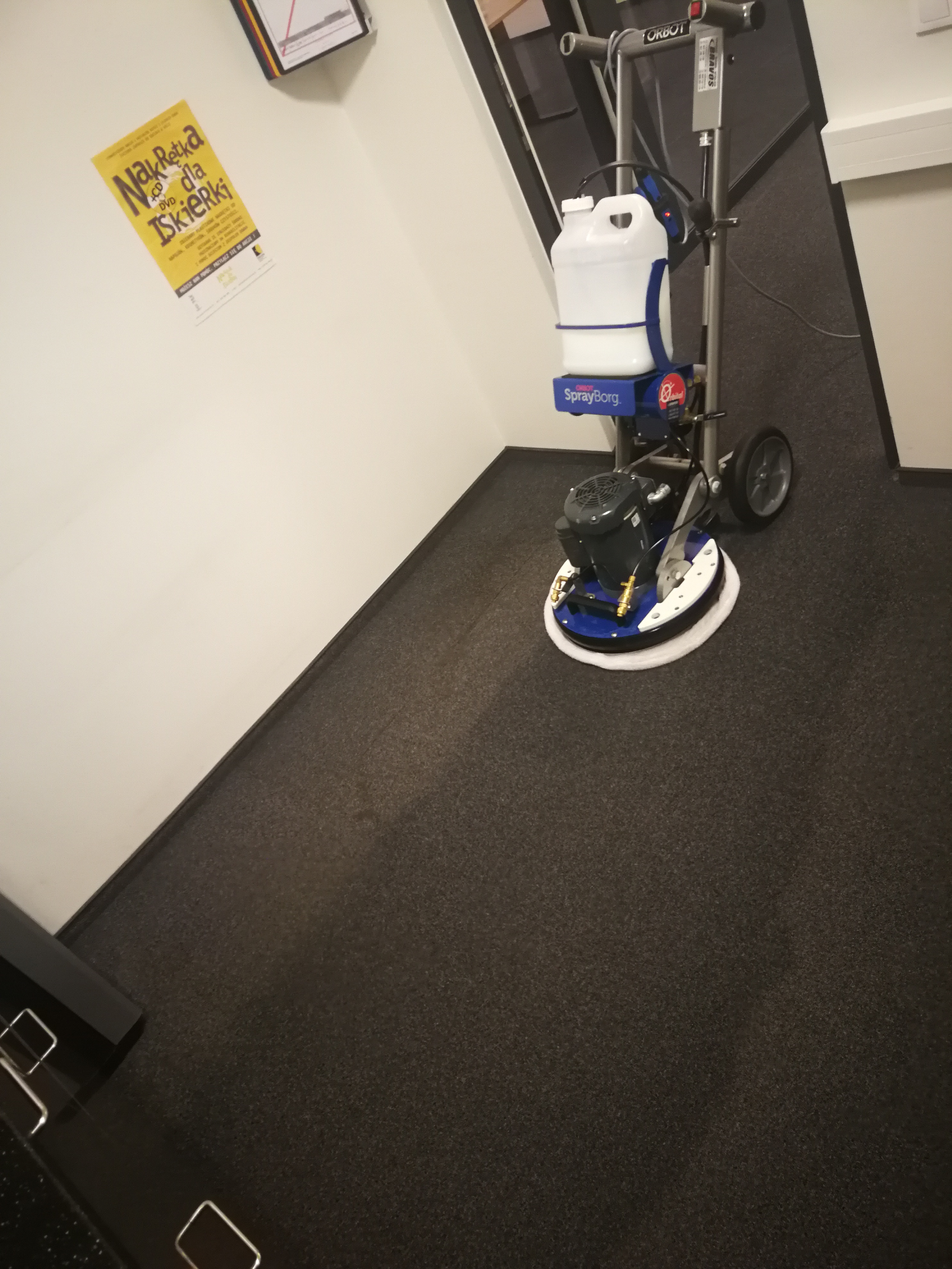 Profesjonalna maszyna czyszcząca Orbot SprayBorg na ciemnoszarym dywanie w biurze, widoczny biały pojemnik na płyn i wentylator silnika.